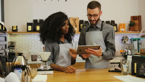 joven barista caucásica mostrando a su compañero de trabajo afroamericano el nuevo menú de café en una tableta durante la jornada laboral