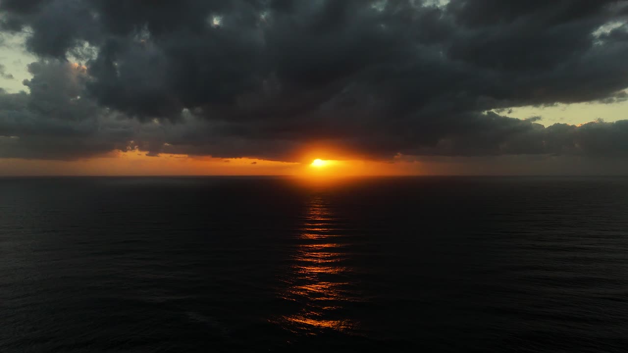 Intense sunset over dark sea horizon with glowing clouds and dramatic sky, aerial dolly along glistening orange light on water