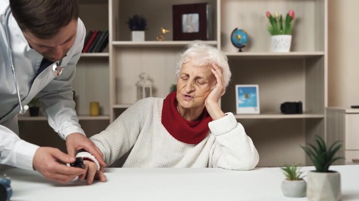 An elderly woman is seated at a table, feeling unwell. Beside her stands a doctor who is attempting to provide her with assistance and measuring her pulse.