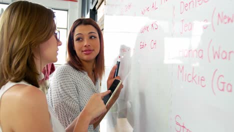 Business-executive-using-mobile-phone-while-colleague-writing-on-white-board