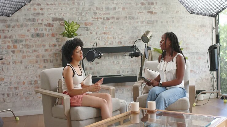 African American women host, guest hosting studio podcast using mics, host asking guest lifting mug