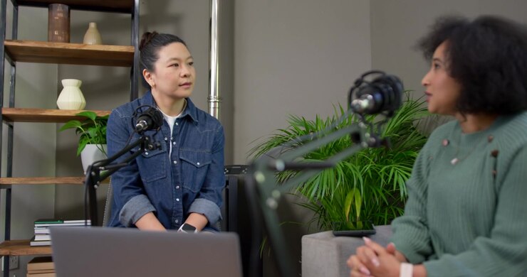 Asian, African American women recording podcast after left prompt in studio, using laptop and mics