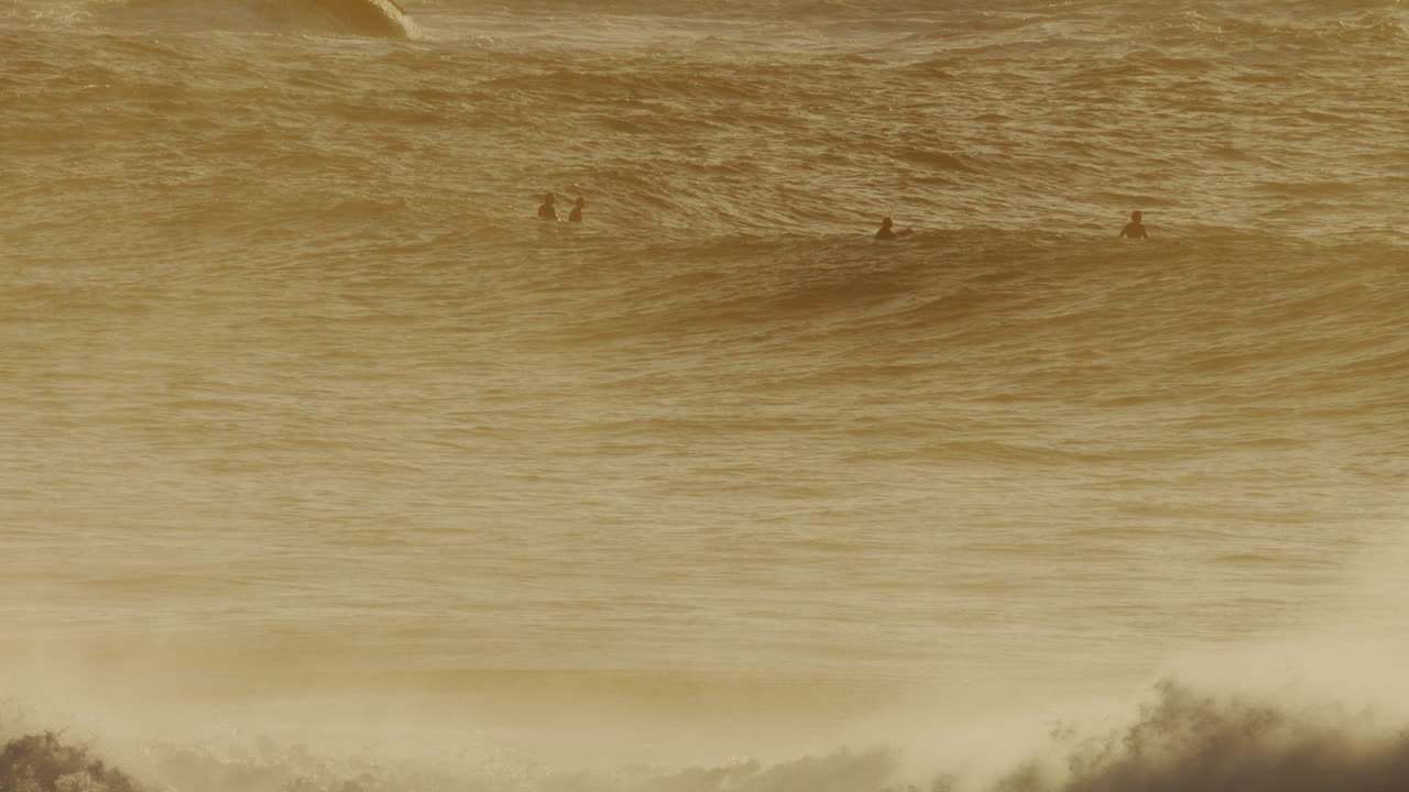 Massive wave crests under hazy gold sky with surfers waiting in lineup on distance