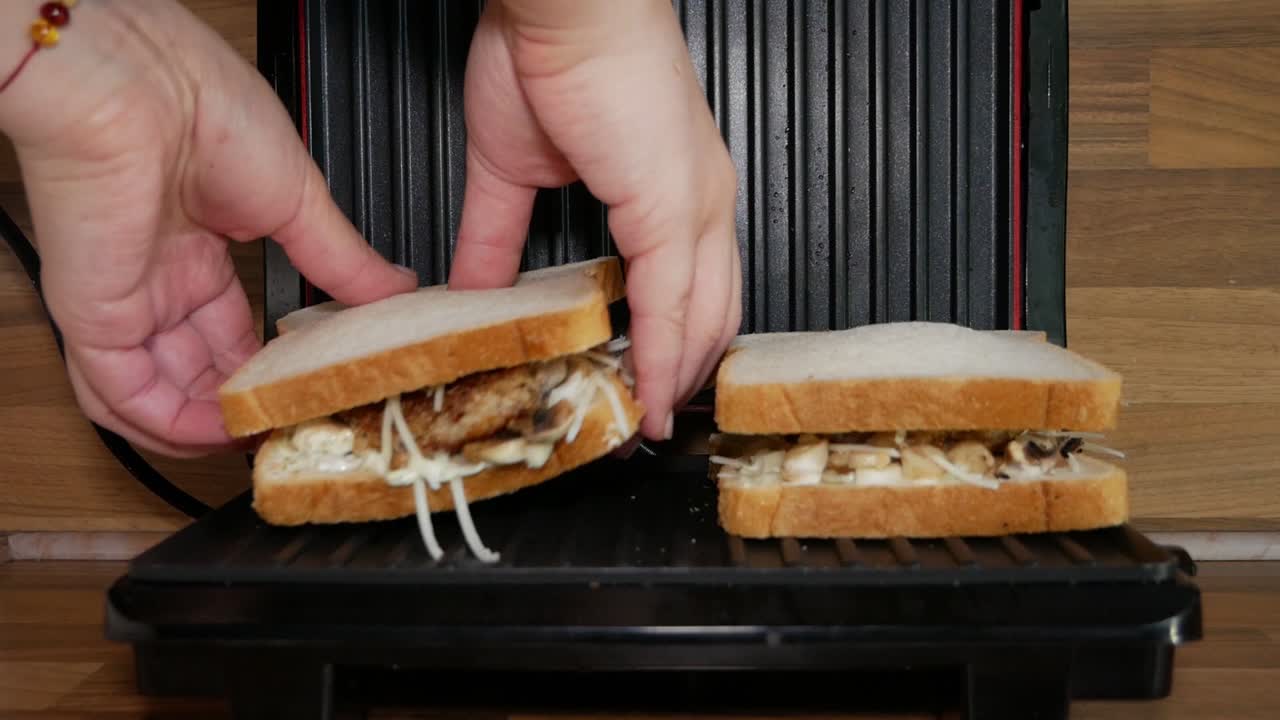 Female home cook putting a second delicious sandwich into electric sandwich maker machine with dipping cheese. Cooking dinner.