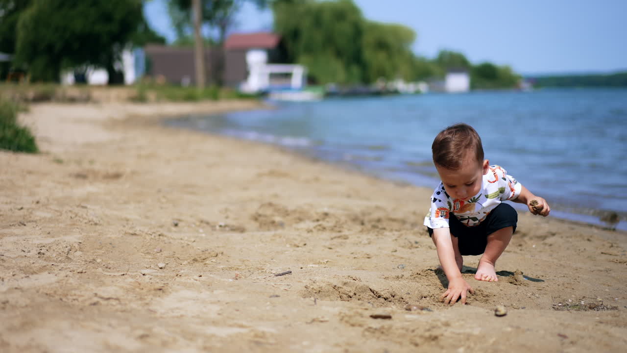 Barefoot Caucasian baby plays peacefully on the river bank. Little boy is interested in wet sand. Blurred backdrop.