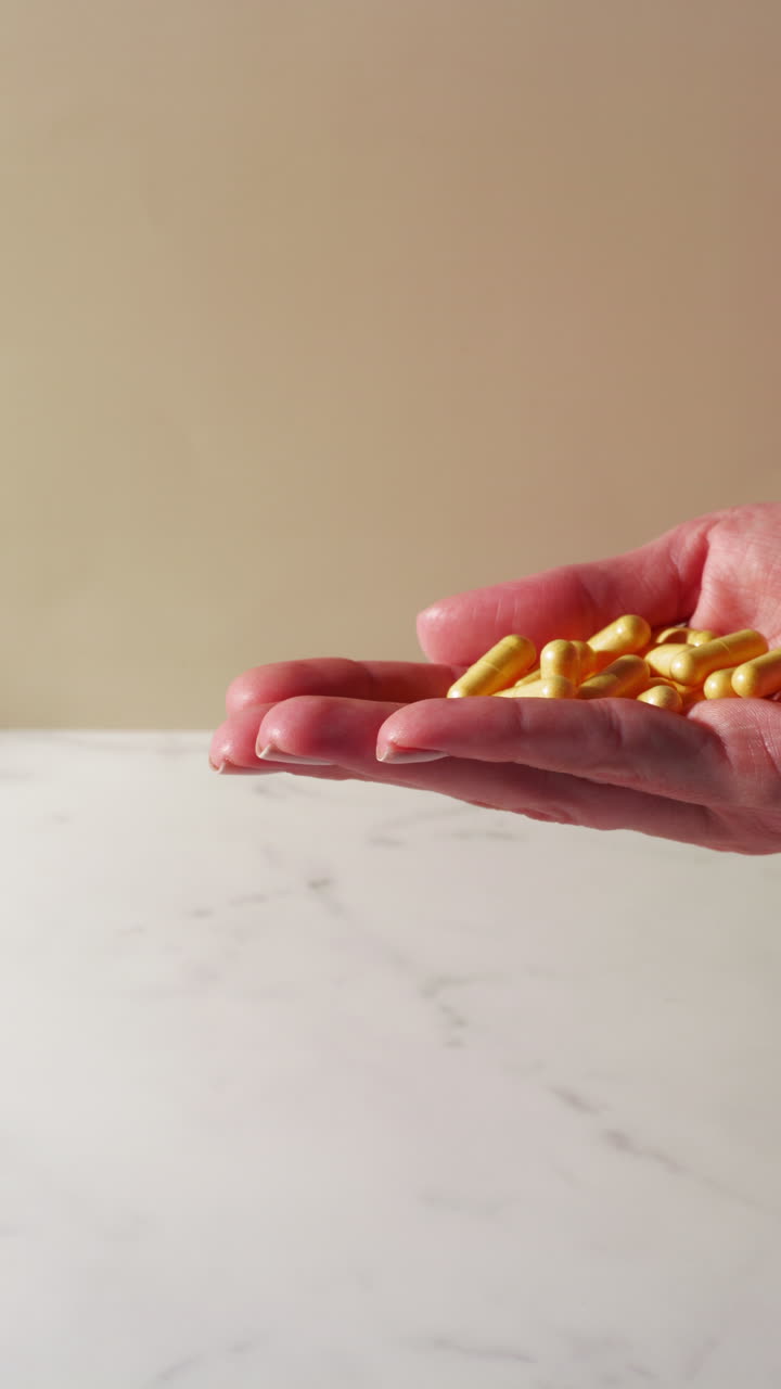 A gentle stream of yellow capsules—possibly vitamin B complex, choline, or cumin—drops from an open hand onto a white marble surface in soft natural light, captured in mid-motion.