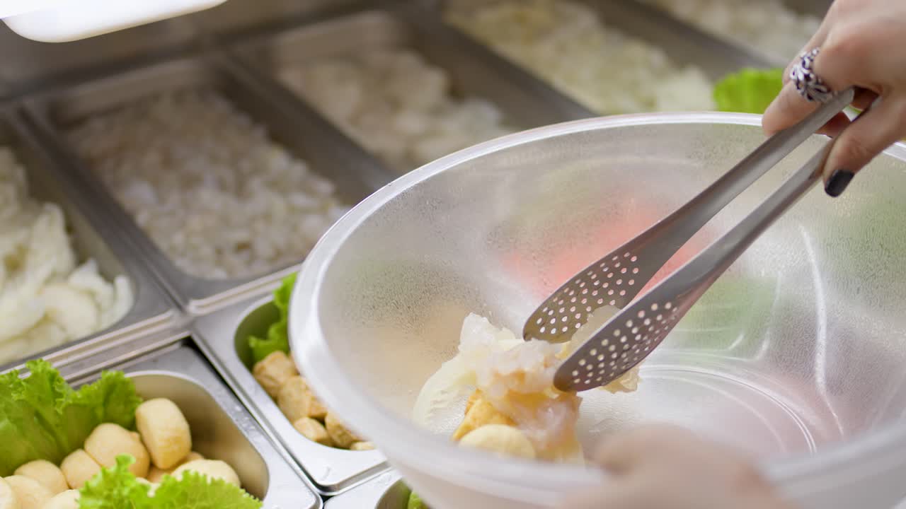 Person uses tongs to pick seafood and vegetables from buffet trays into a clear bowl