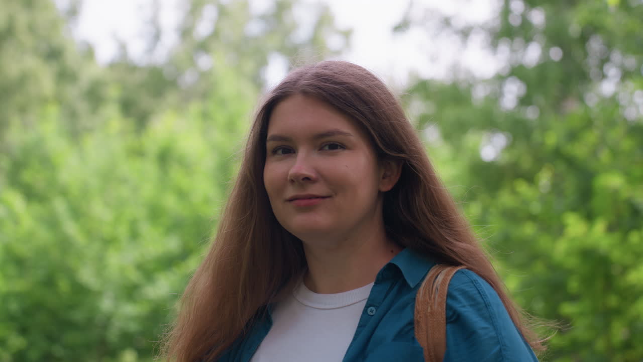 Una mujer preciosa sonríe con confianza en un exuberante parque verde, vestida con una camisa azul y una camiseta blanca, de pie con calma bajo la luz del sol, mostrando un encanto natural y energía positiva