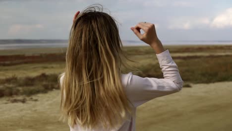 relaxed woman is dancing alone standing on sand beach with closed eyes, water front on background