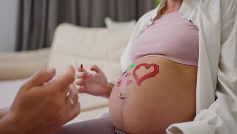 Close-up-a-happy-man-uses-a-palette-to-paint-on-the-belly-of-his-pregnant-wife-during-their-leisure-time-and-waiting-for-the-baby-at-home