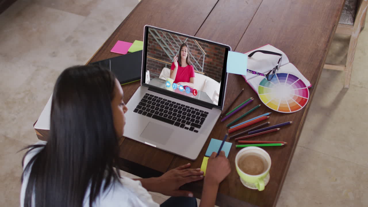Mujer caucásica usando una laptop en videollamada con una colega, tomando notas
