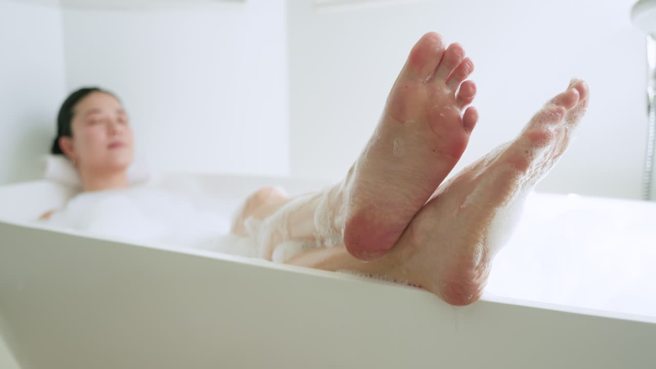 Mujer recostada en la bañera, levantando las piernas y mostrando las plantas de los pies, mientras la cámara enfoca para el bienestar.