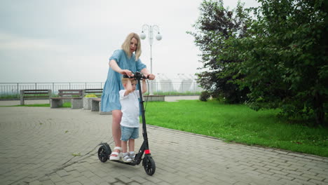 a mother and her young son stand together on an electric scooter, on an interlocked pathway