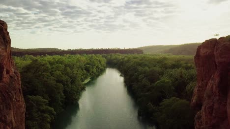 Drone-flying-straight-out-of-a-gorge-between-two-large-red-rock-walls-following-a-river-surrounded-by-green-trees-and-vegetation