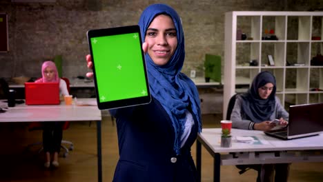 beautiful smile of arabic female who shows green screen on tablet and stands in brick office among her arabian colleagues, indoor illustration, modern life