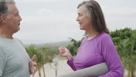 Happy-senior-caucasian-couple-holding-yoga-mat-and-talking-on-beach,-in-slow-motion