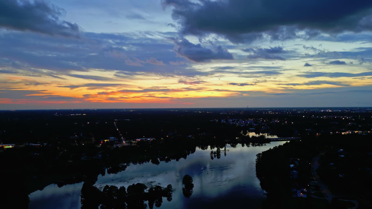 A dramatic sunset that's slowly fading away over a dark reflective river with forested shoreline, colorful clouds and fading light, aerial shot flying forward
