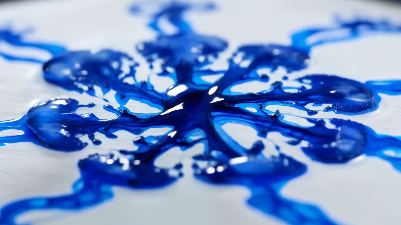 Dispensing nozzle releasing blue liquid on white dish, drop making ripples, forming flower pattern