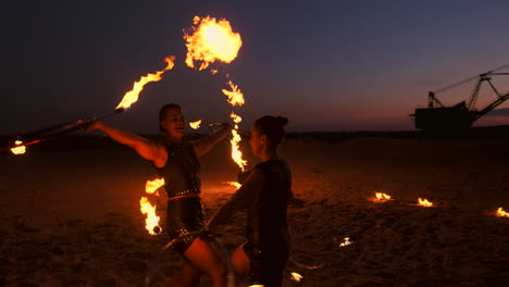 Eine-Gruppe-Von-Männern-Und-Frauen-Zeigt-Nachts-Eine-Feuershow-Auf-Dem-Sand-Vor-Dem-Hintergrund-Von-Feuer-Und-Turmdrehkränen.