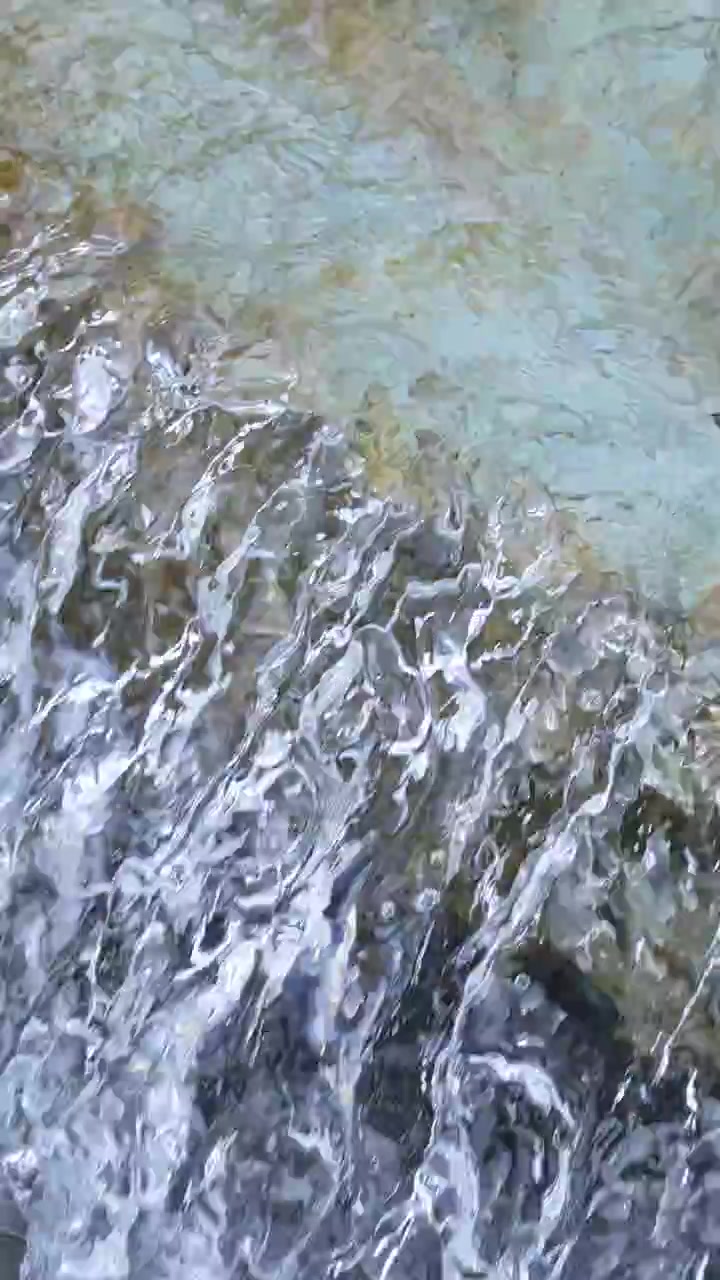 Primo piano di acqua trasparente di ruscello che scorre impetuosa su rocce lisce alla luce naturale del giorno, con una telecamera stabile che cattura texture superficiali dettagliate e il movimento fluido.
