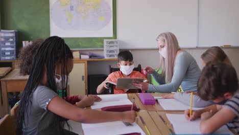 Female-teacher-wearing-face-mask-teaching-disable-boy-by-using-digital-tablet-at-school