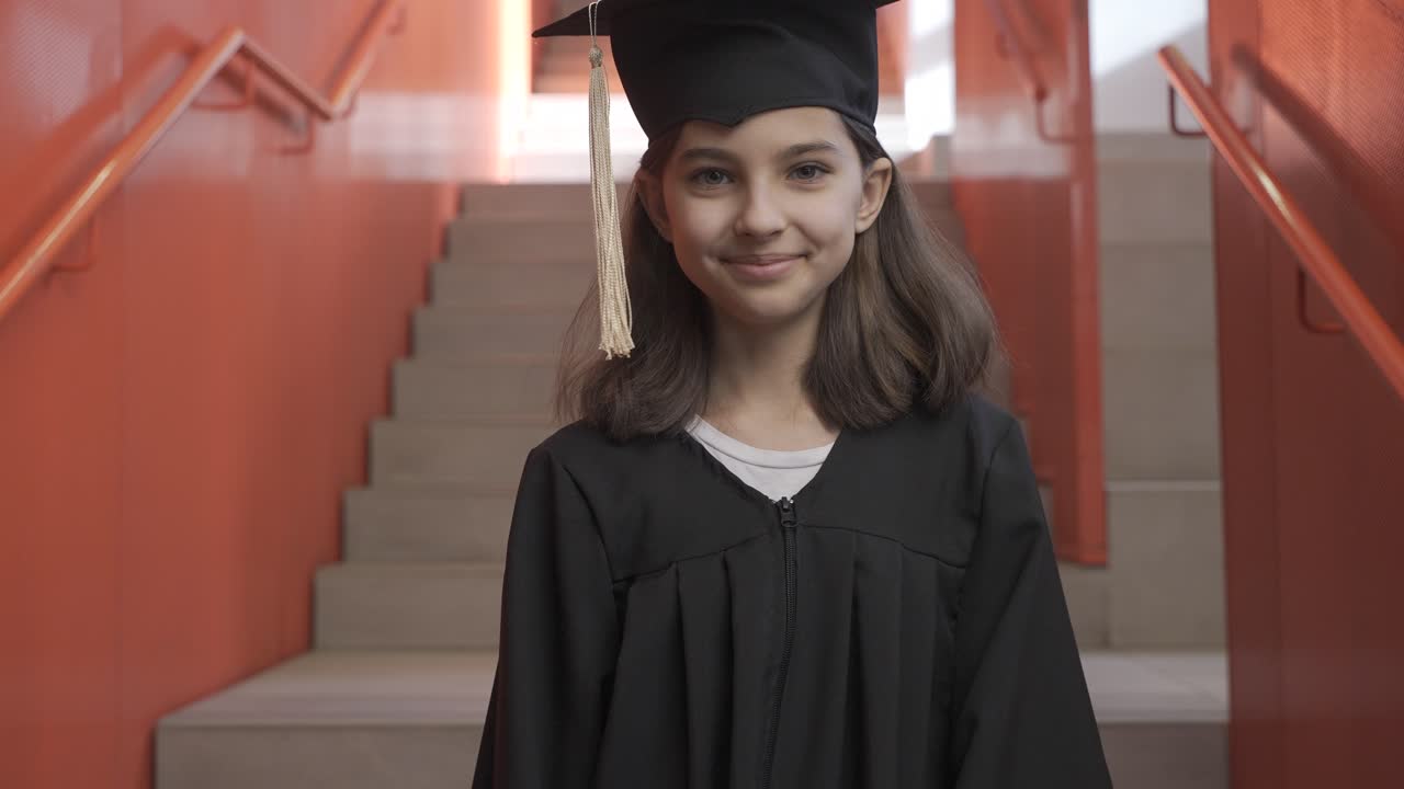 Portrait of a happy preschool female student in cap and gown going down the stairs, holding graduation diploma and looking at the camera
