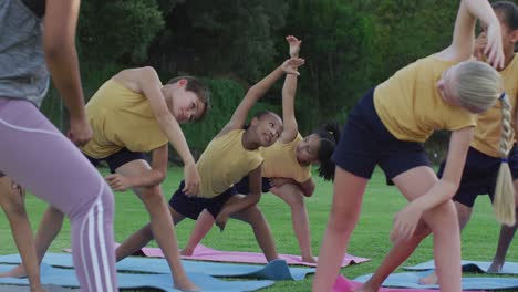 mixed race female teacher showing diverse group of schoolchildren yoga stretching exercises outdoors