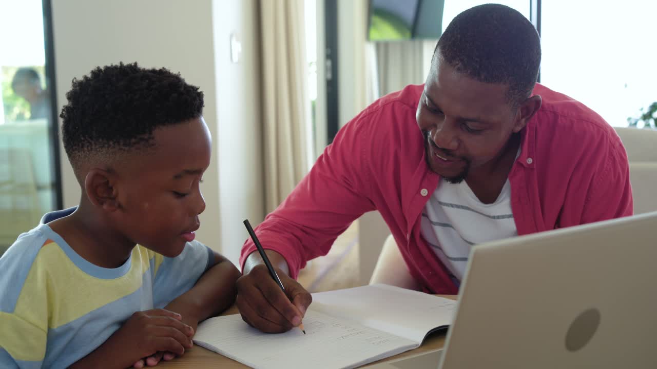 African American father and child at table, father writing book, giving pencil, child trying answer