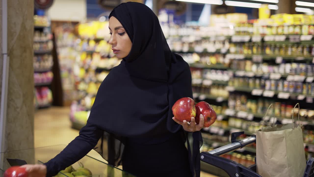 mujer musulmana comprando comestibles, tomando manzanas rojas del pasillo de frutas
