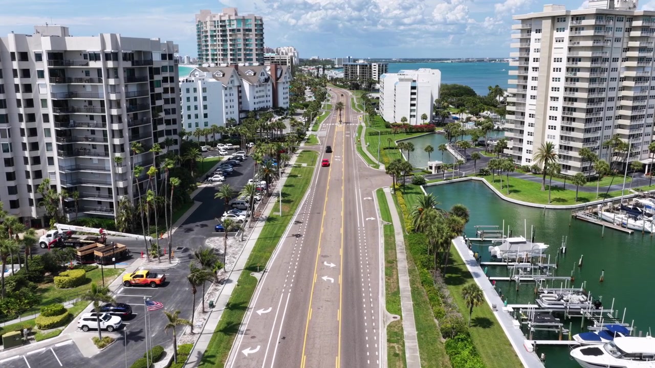 Viale alberato di palme si estende tra condomini sul lungomare e un porto turistico a Clearwater Beach, con veicoli in movimento, barche ormeggiate, un canale e le acque del Golfo sullo sfondo. Florida, USA. Ripresa aerea con drone
