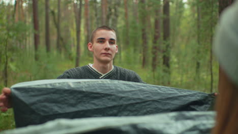 young couple unpack red gray tent on forest floor with boyfriend leaning forward to kiss partner then stepping back with calm expression, both handling tent canvas amid green woodland