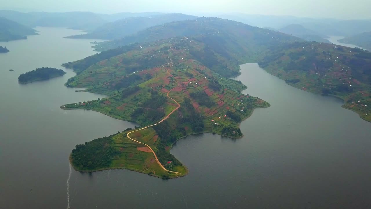 Scenic drone shot shows Lake Bunyonyi’s peninsula surrounded by hills and patchwork farmlands, revealing the lake’s intricate shoreline and Uganda’s lush highland terrain from above
