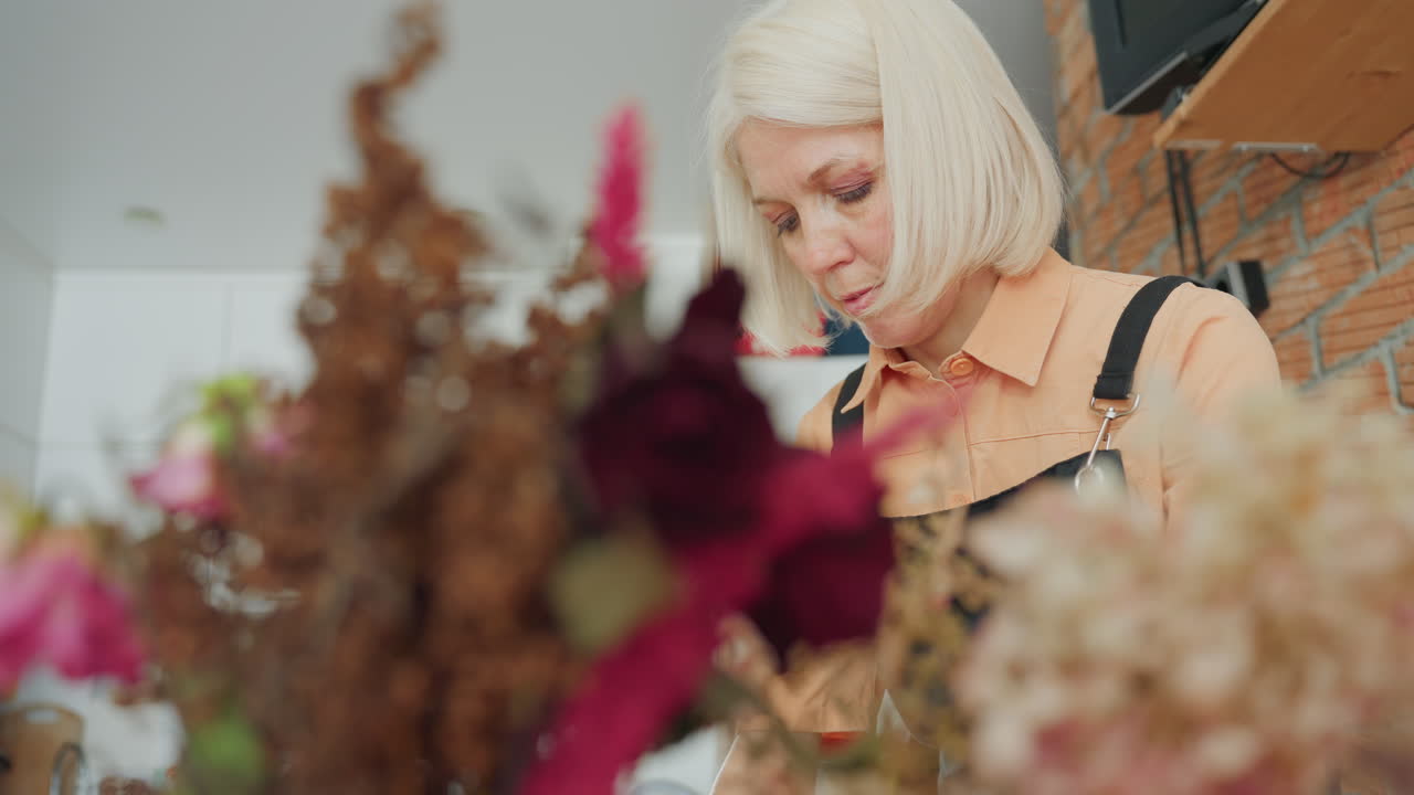 Senior woman florist in apron focusing on floral arrangement, blurred dried and fresh flowers in foreground, creative workspace atmosphere showing concentration, during flower composition work