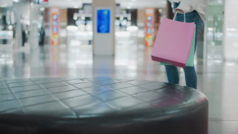 partial view of lady in white outfit walking toward leather seat with shopping bags in hand, placing pink and mint bags on leather seat in shopping mall, blurred background with modern retail setting