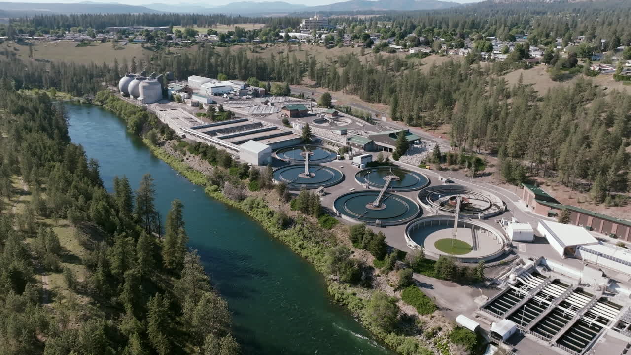 A wide aerial shot of the Spokane water treatment facility showcases the entire plant layout along the scenic Spokane River, surrounded by forested hills and residential areas.