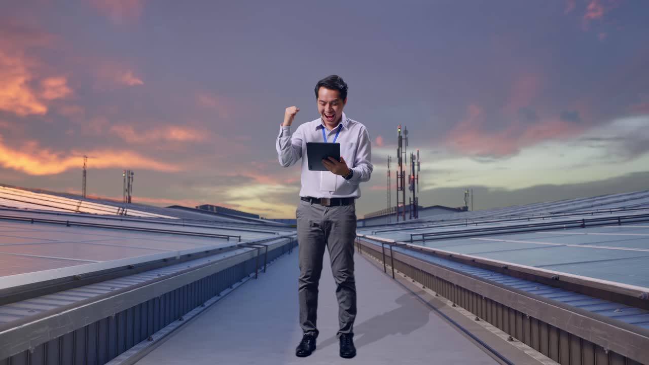 Un professionista asiatico, a figura intera, in piedi con il suo tablet, ispeziona pannelli solari industriali sul tetto; alza il pugno in segno di esultanza dopo aver digitato sul tablet