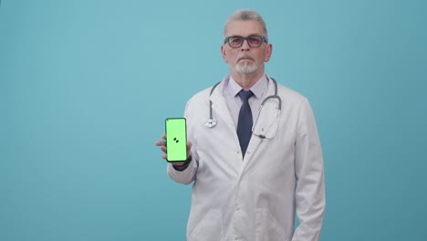 serious adult man doctor holds a phone with an app in the studio on a blue background. mockup greenscreen