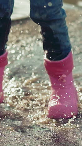 closeup slow motion little child runs and jumps into deep puddle in rainboots and jeans with all over span, picking up large splash fountain, soiling clothes, and wet pants, in sunny weather