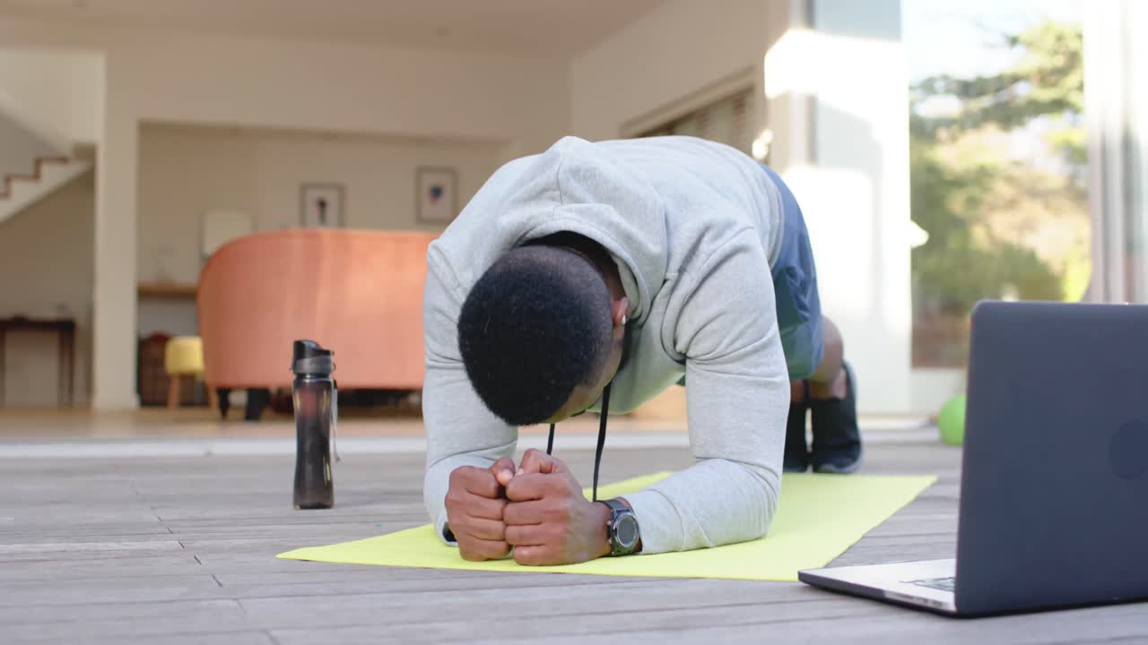 Focused african american man exercising on mat with laptop at home, slow motion