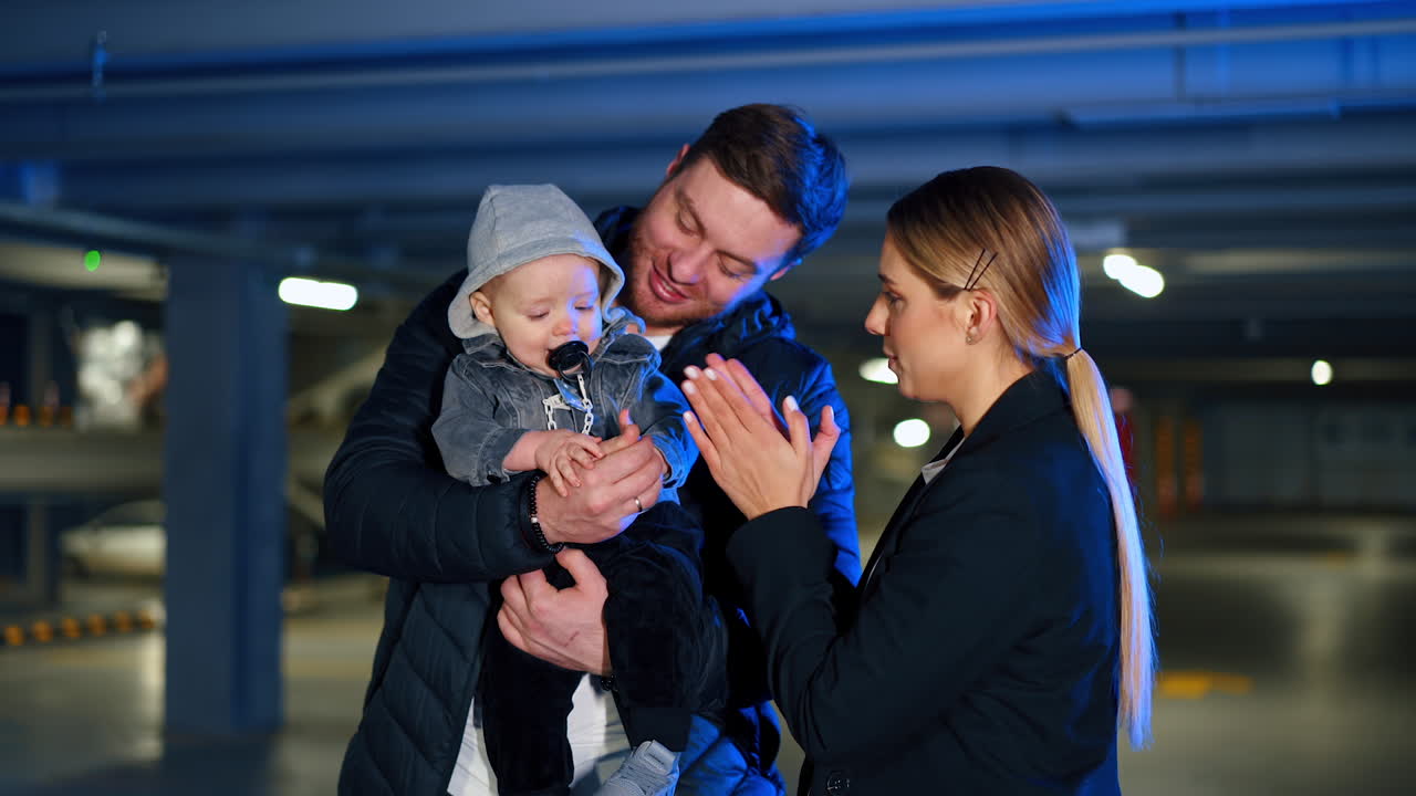 Bearded man holding a cute infant baby with pacifier in mouth. Happy mom claps her hands to entertain her child.