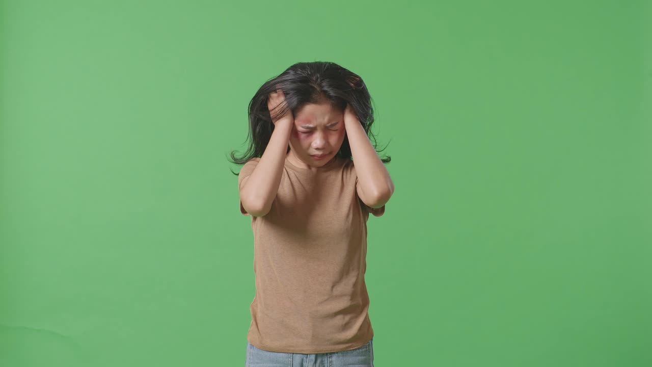 Young Asian Woman Victim Of Violence With Bruise On Face Being Stressed And Shaking Her Head Having A Headache On Green Screen Background In The Studio