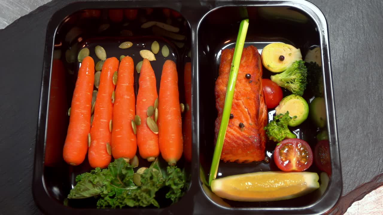 A top-down view of a healthy, pre-packaged meal prep or food delivery container with grilled salmon, fresh vegetables, and carrots for a nutritious diet
