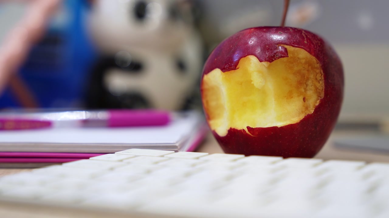 Extreme close up of a bitten red apple on a modern office desk during bright daylight. Cinematic shallow depth of field captures a blurred white keyboard and pen. Perfect for workplace wellness.