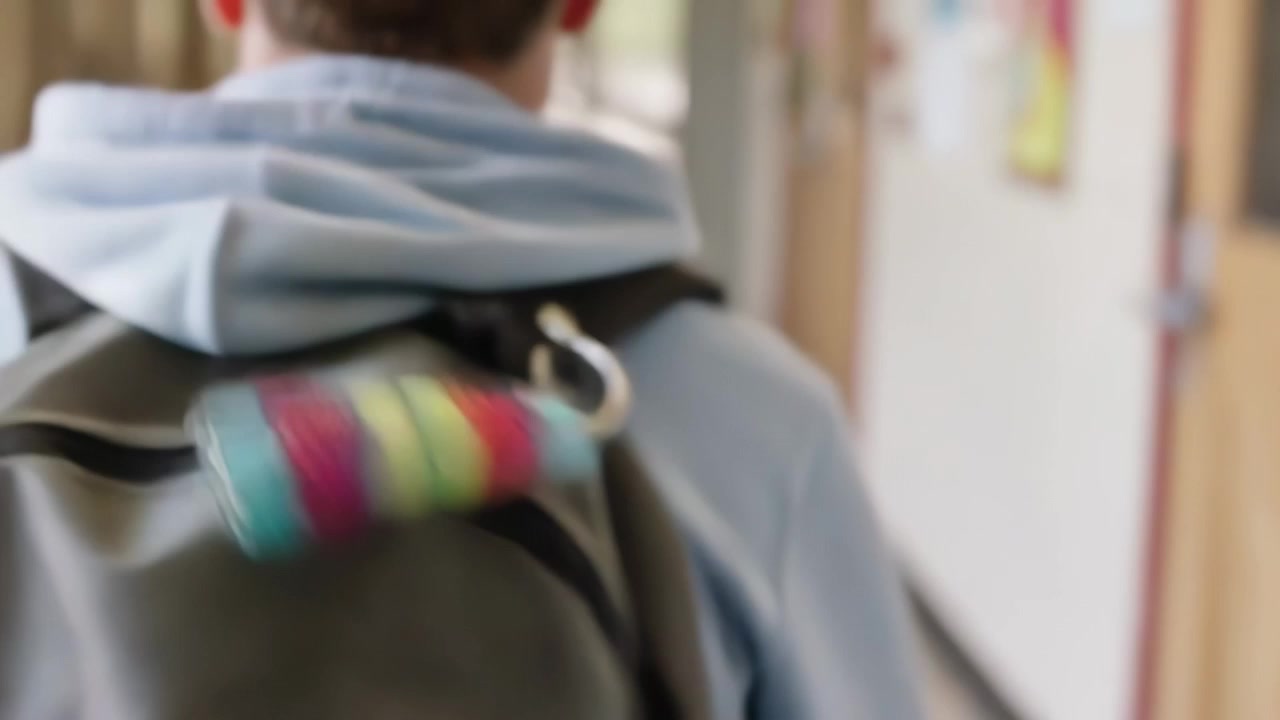 Hoodie student walking to class in school hall, gray-backpack pom-pom swinging, camera pulling back