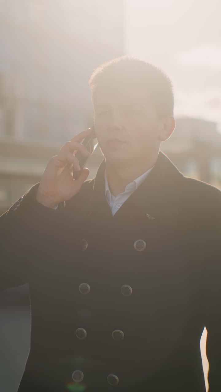 검은 코트를 입은 은행가는 차를 떠나 심각한 표정으로 주머니에서 전화를 꺼내서 전화에 응답합니다. 주차된 자동차와 현대적인 건물이 있는 활기찬 도시 지역에서