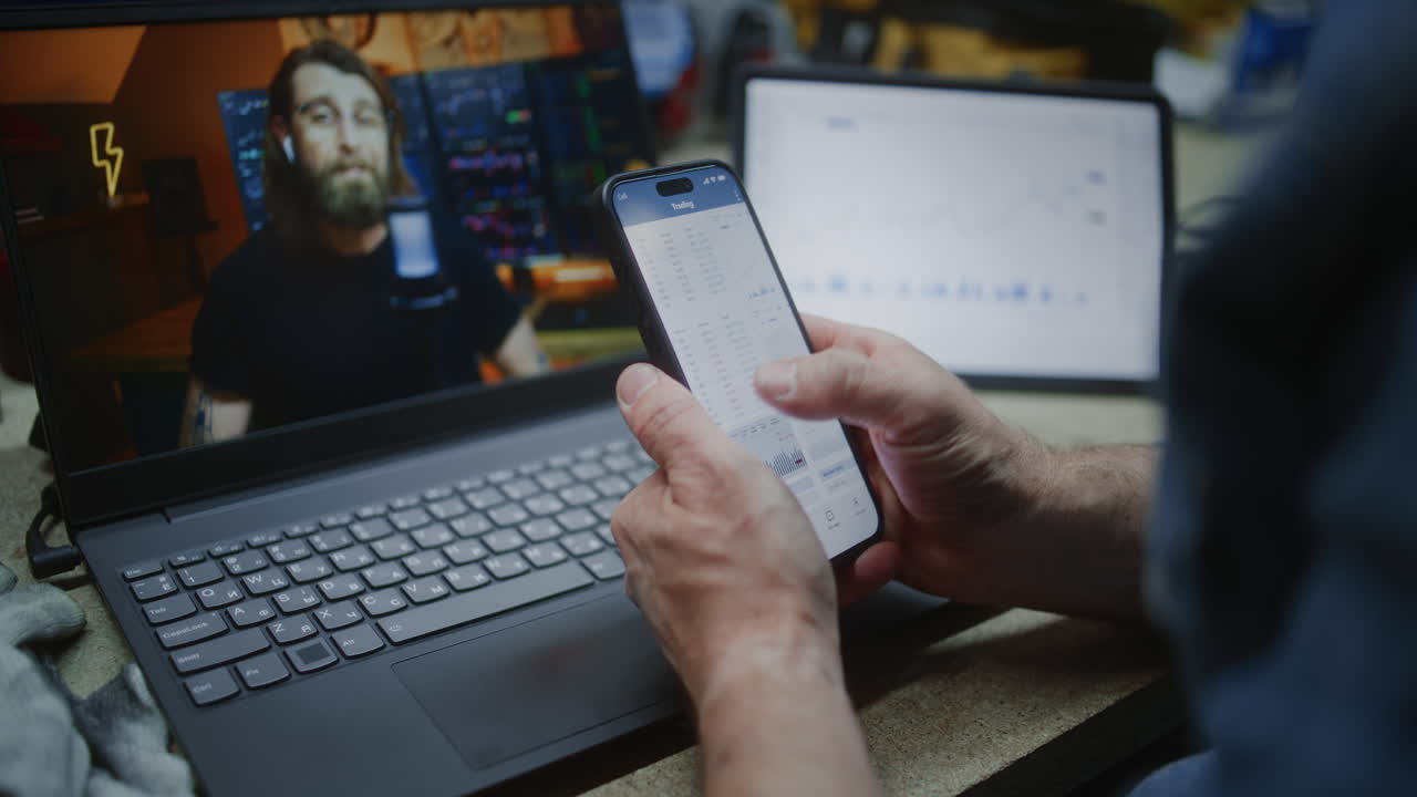 Worker Watching Online Trading Education on Laptop, Monitoring Real-Time Stocks and Exchange Market Charts Using Smartphone. Man Remote Learning Trading During Working Day in Workshop. Vertical Shot.