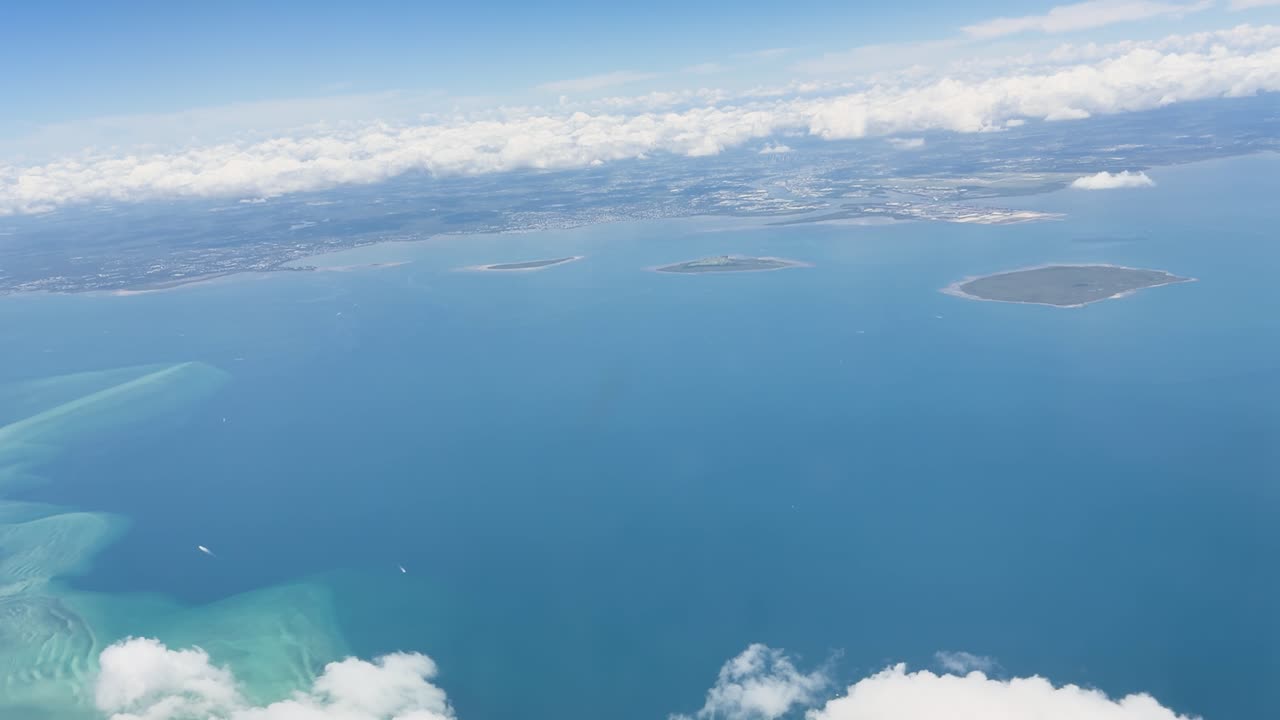 熱帯の海岸線、小さな島々、ターコイズブルーの海、雲、そして水平線が広がる、飛行機の窓から見えるなめらかな景色