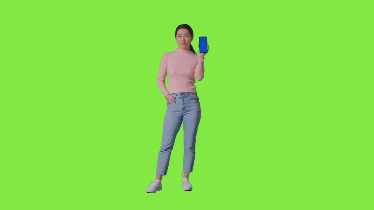Ritratto in studio a figura intera di donna che tiene un cellulare con schermo blu verso la fotocamera, su sfondo verde 1.
