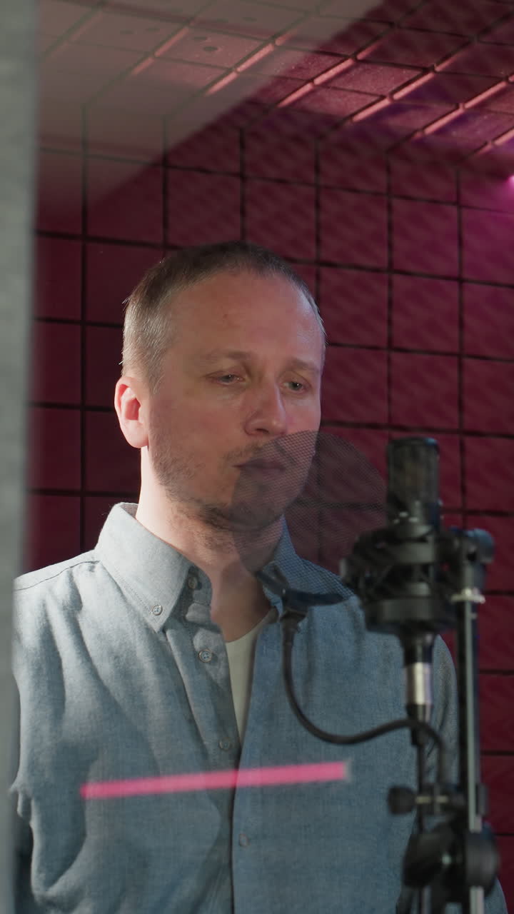 un hombre con una camisa de manga larga azul se para frente a un micrófono de estudio con una cara recta, mientras extiende su mano en una habitación a prueba de sonido roja