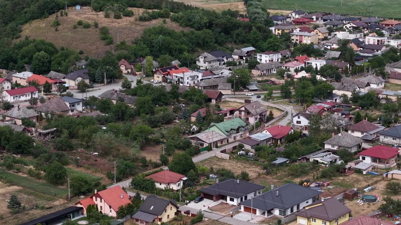 An aerial view of a residential village, showcasing scattered houses amidst lush green trees and rolling hills under daytime lighting.